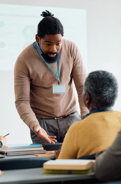 Black teacher talking to senior man during adult education training class in lecture hall.